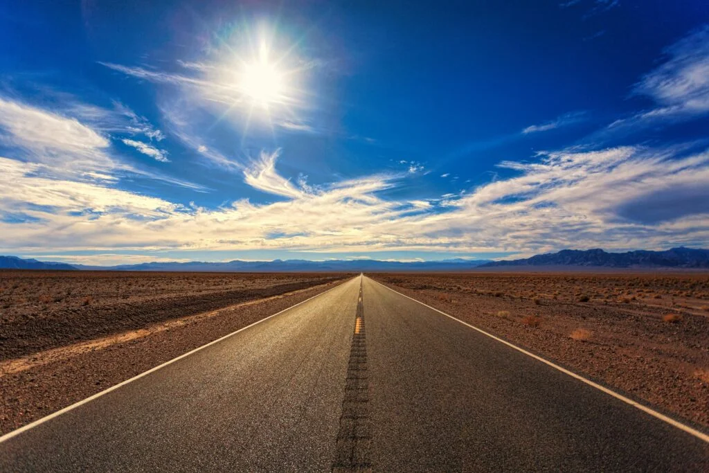 A long, straight road stretches across a dry desert landscape under a bright blue sky and shining sun.